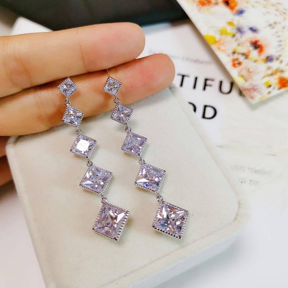 Silver earrings with square-shaped stones held by a hand on a textured surface.