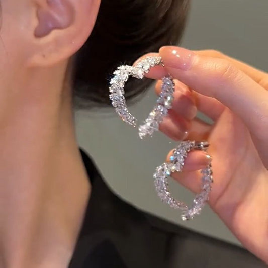 Person holding a pair of diamond hoop earrings close to their ear.