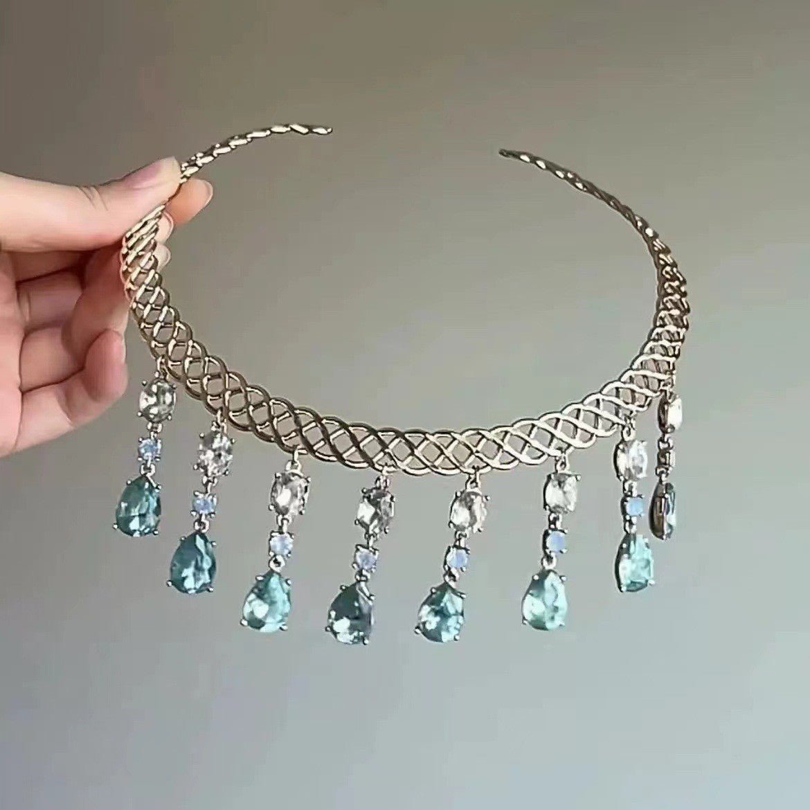 Gold necklace with green teardrop pendants held by a hand against a neutral background