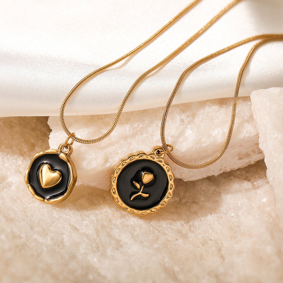 Two gold necklaces with black pendants featuring heart and flower designs on a textured beige surface.