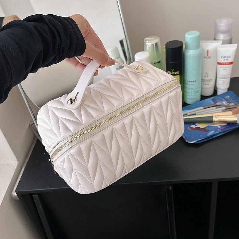 White quilted cosmetic bag held by a person with beauty products on a table in the background.