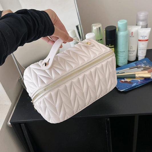 White quilted cosmetic bag held by a person with beauty products on a table in the background.