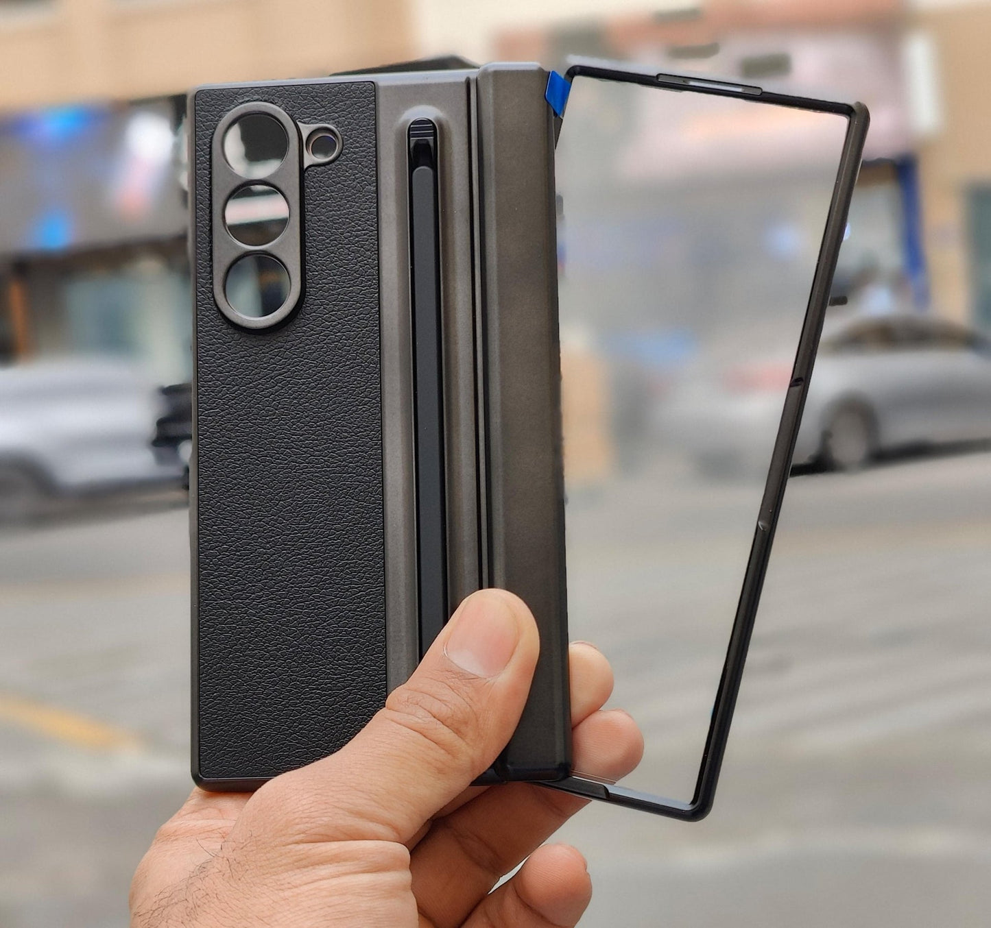 Person holding a black phone case with a reflective surface, blurred city street in the background