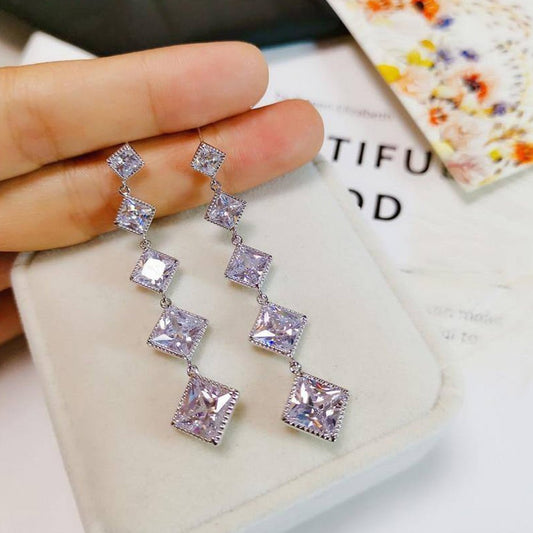 Silver earrings with square-shaped stones held by a hand on a textured surface.
